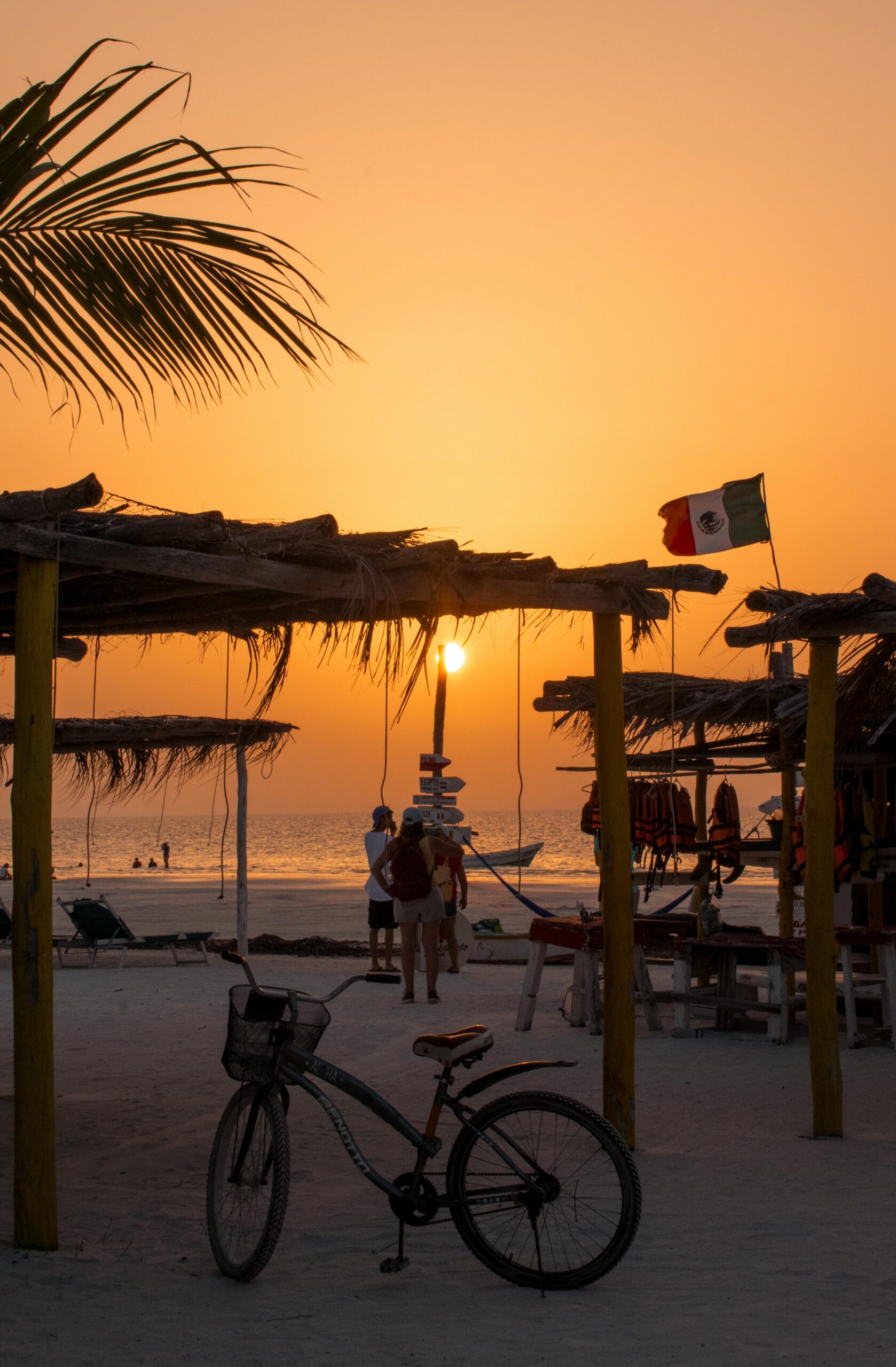 Sunset on a Mexican beach with palm trees, huts, and a bicycle, Mexican flag waving