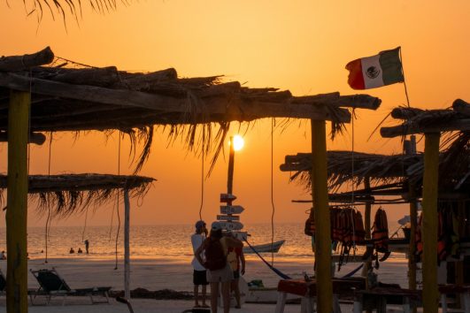 Sunset on a Mexican beach with palm trees, huts, and a bicycle, Mexican flag waving